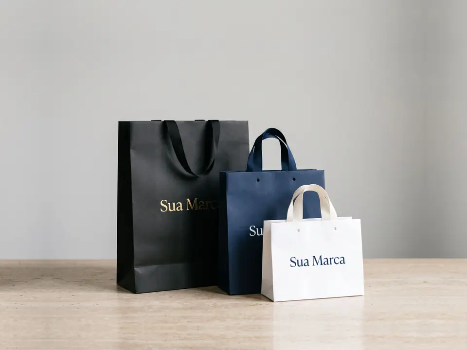 Três sacolas de papel personalizadas, uma preta, uma azul-marinho e uma branca, com a marca impressa, sobre uma mesa.