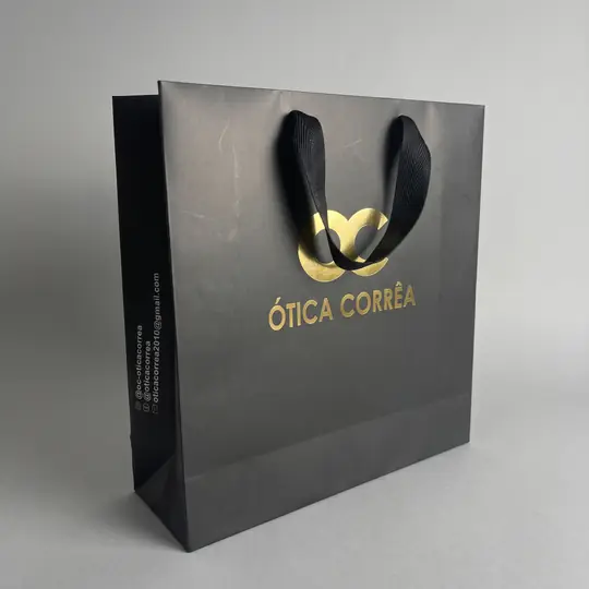 Sacola de papel preta com a logo em dourado e alça de fita preta